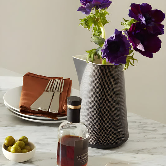 Clear shot glass with drink on marble table with bottle, purple flowers, and olives.