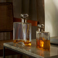 Two glass whiskey decanters with orange liquid on a marble table, next to a chair.