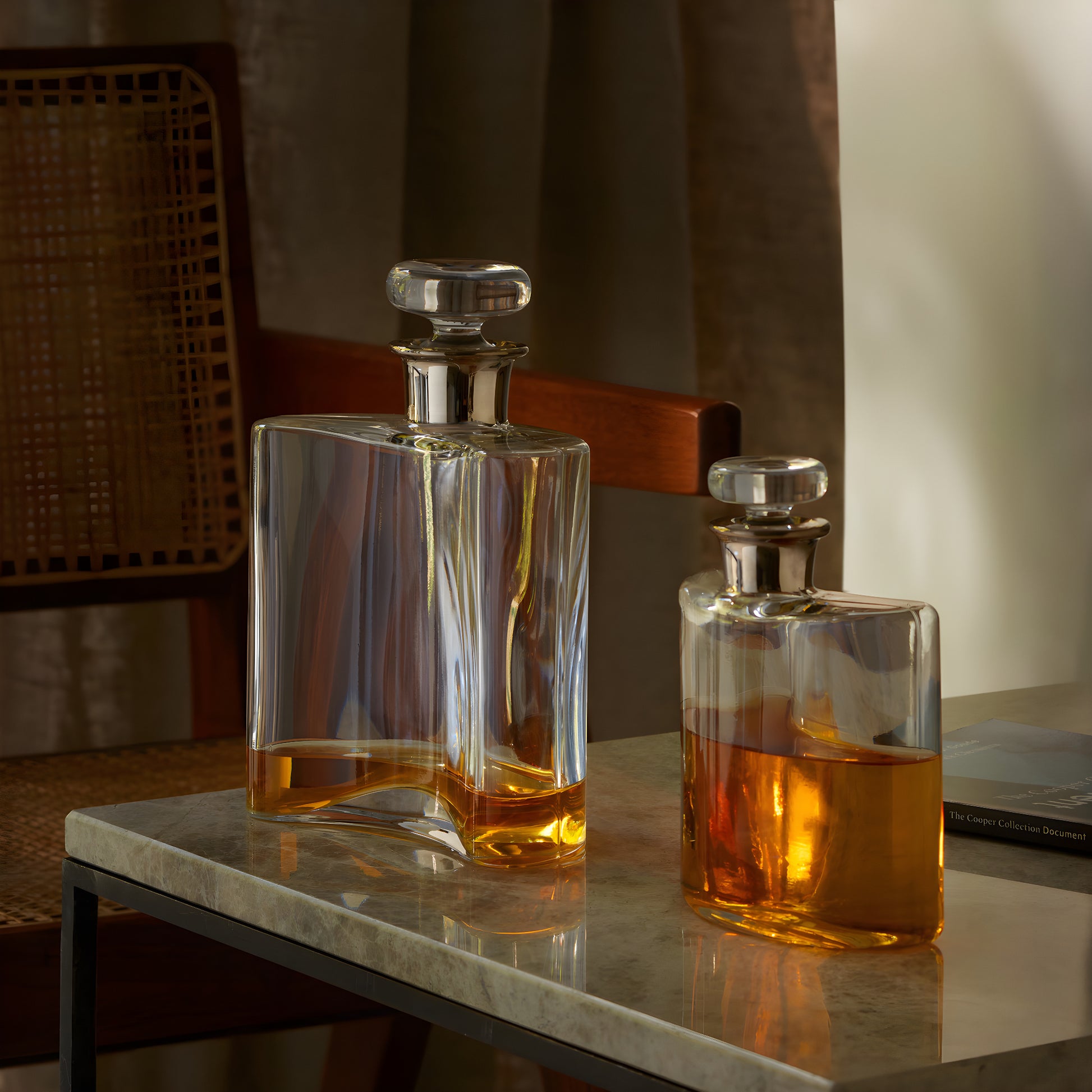 Two glass whiskey decanters with orange liquid on a marble table, next to a chair.