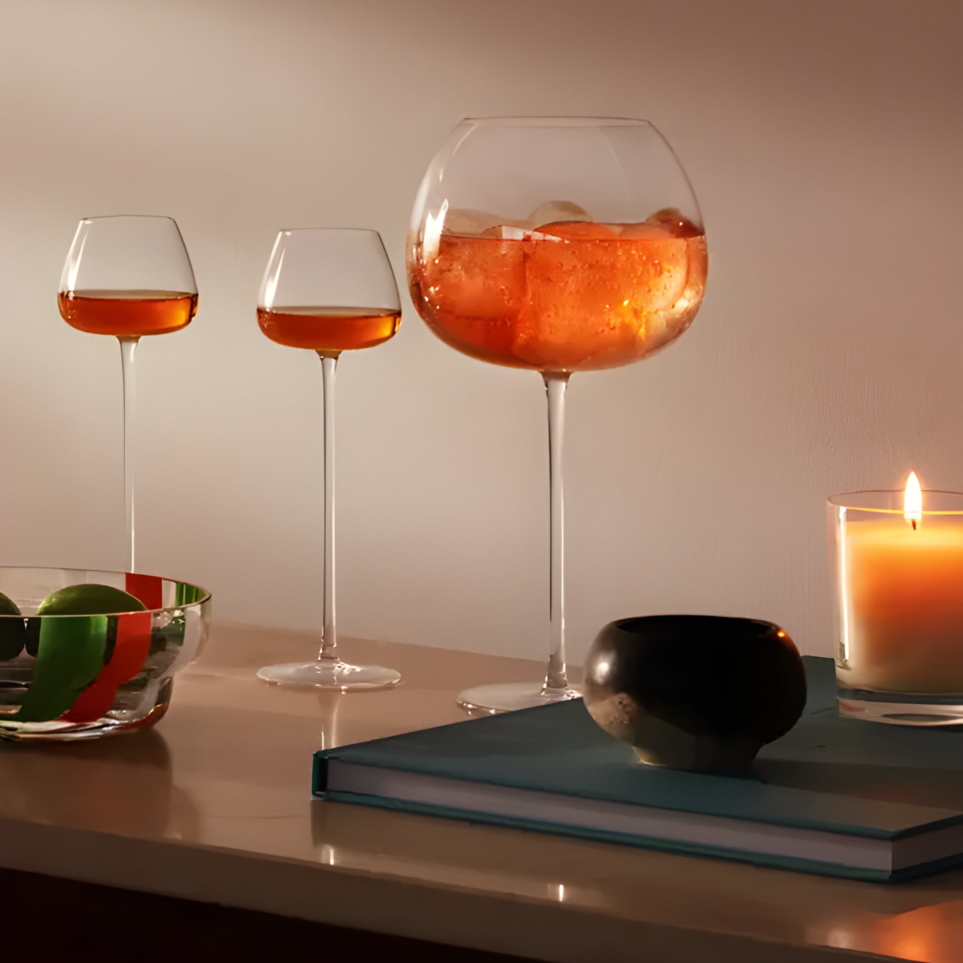 Set of balloon glasses with orange cocktails on candlelit table with books and bowl.
