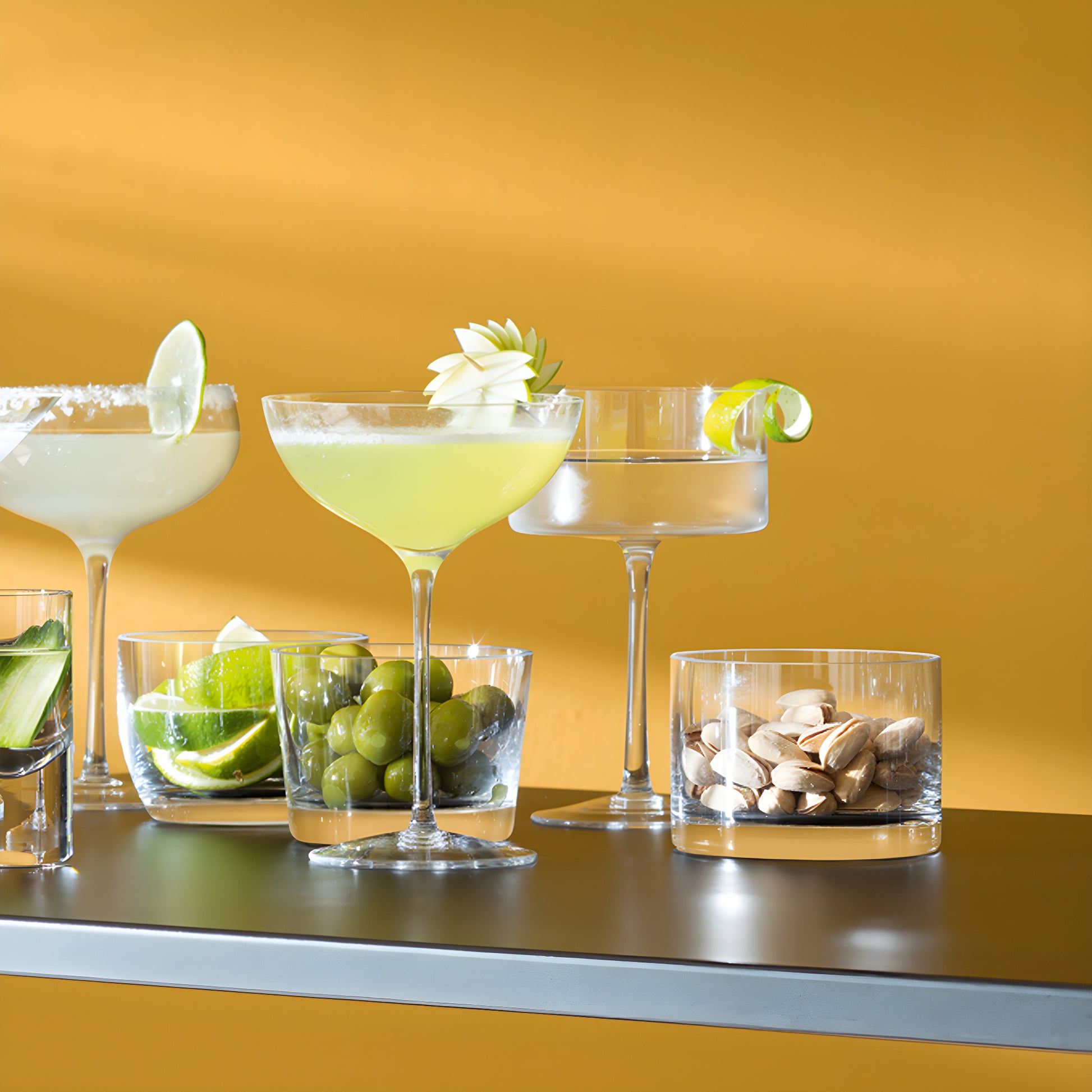 Cocktail glass with apple garnish beside olives and limes on a yellow background.