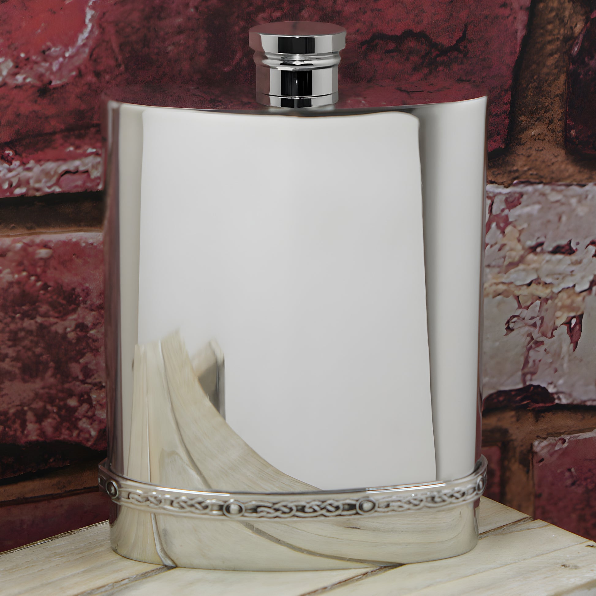Engraved pewter hip flask with Celtic design against brick wall background.
