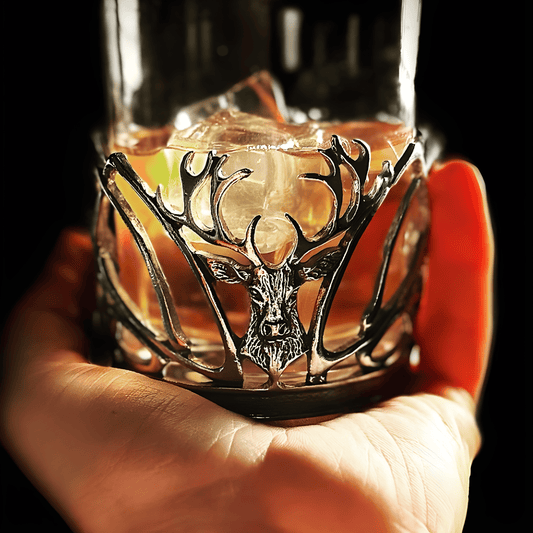 Hand holding a metal-inlaid whisky glass with stag design and ice cubes.