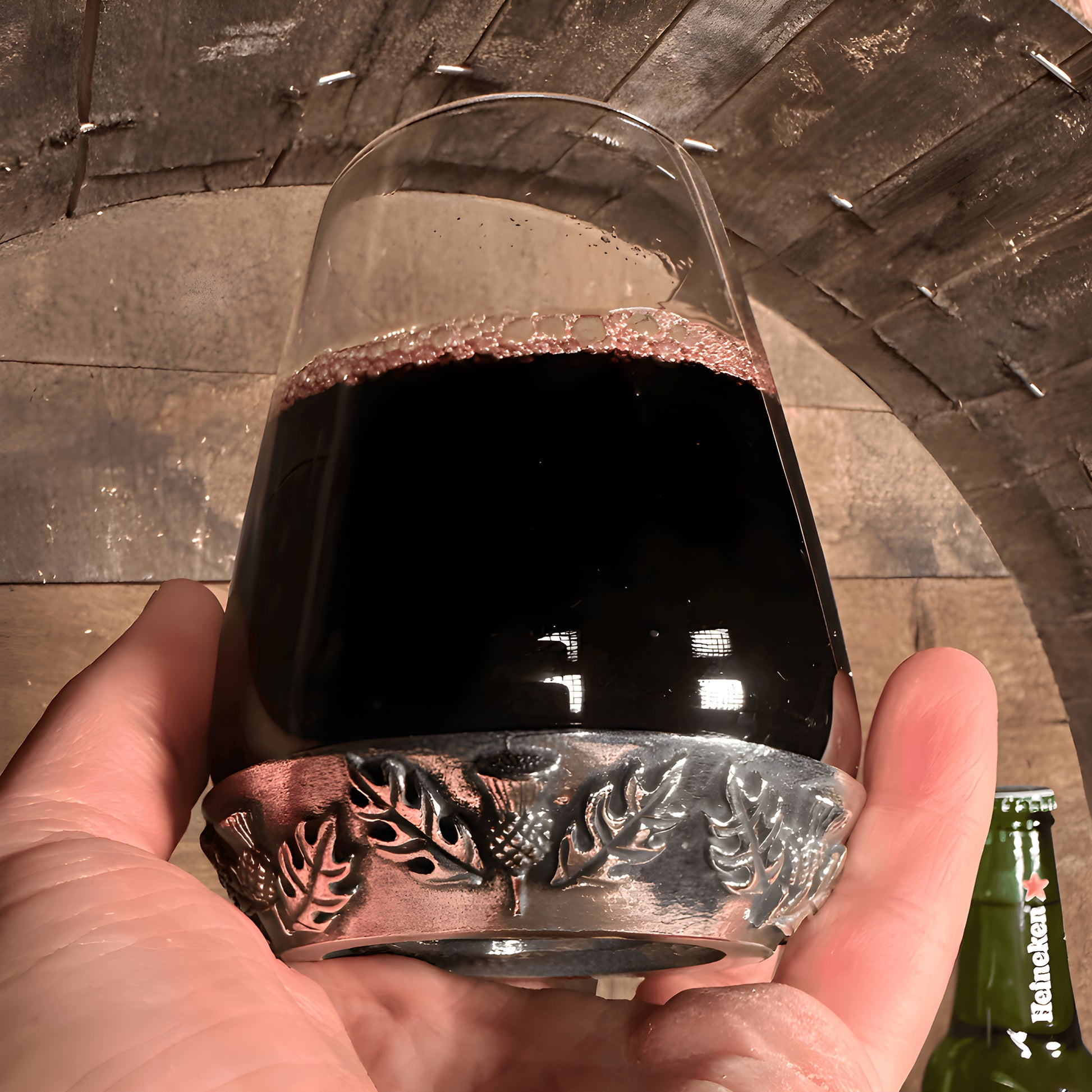 Hand holding a thistle tumbler glass filled with red wine, featuring ornate metalwork in rustic setting.