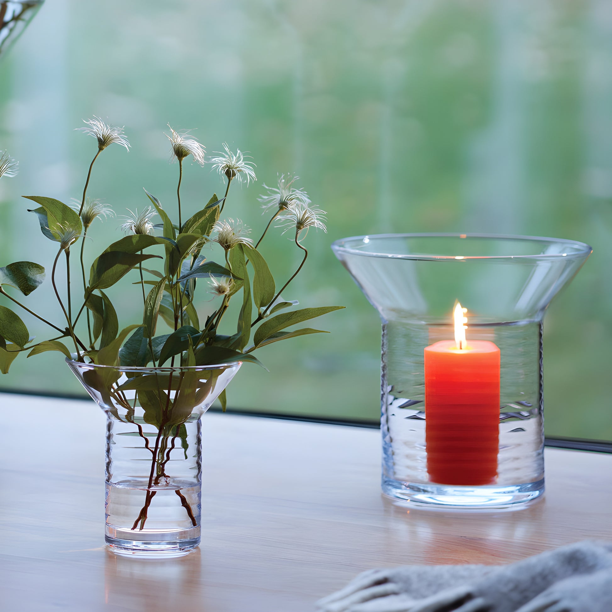 Clear glass vase with red candle and greenery on wooden table by window.