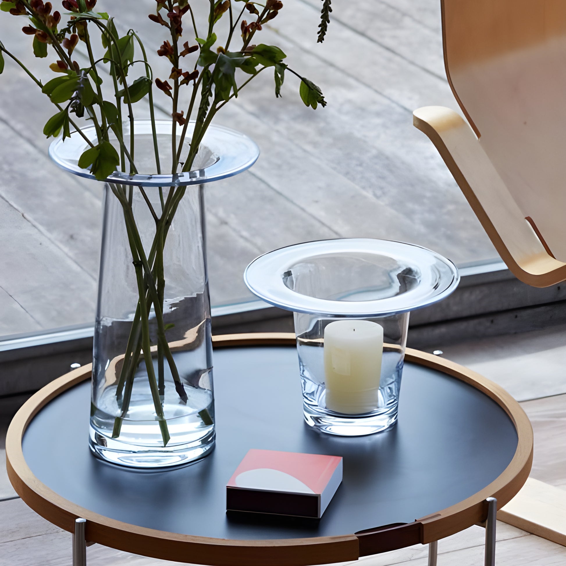 Glass flower vase on a round wooden coffee table with candle and chair nearby.