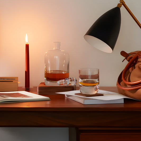 Whisky tumbler set on a wooden desk with a candle, decanter, and lamp.