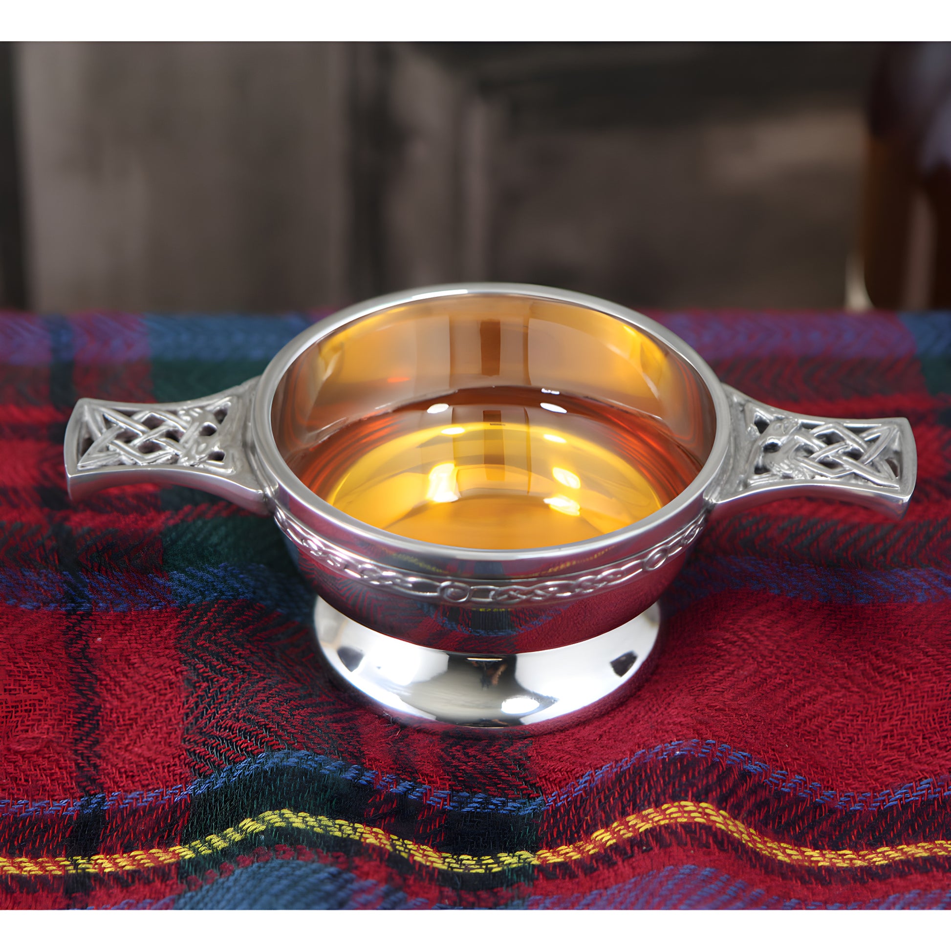 Pewter quaich bowl with Celtic knot handles filled with whiskey on tartan cloth.