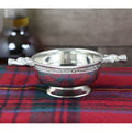 Pewter quaich bowl with Scottish thistle handles on tartan fabric.