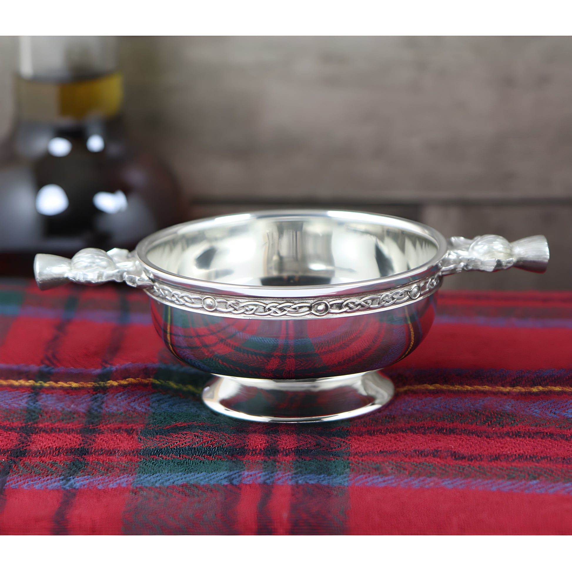 Pewter quaich bowl with Scottish thistle handles on tartan fabric.
