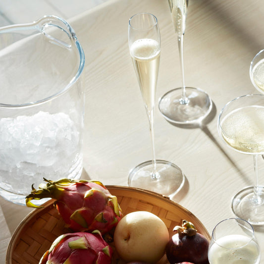 Champagne flutes and coupe glasses with fruit platter by an ice bucket on a bar table.