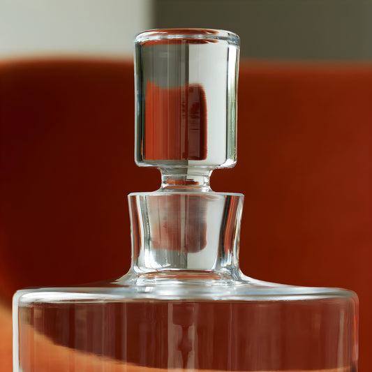 Closeup of a clear glass decanter with stopper against a red background.