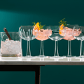 Four gin and tonic glasses filled with ice and garnishes on a white table, with a green wall background.