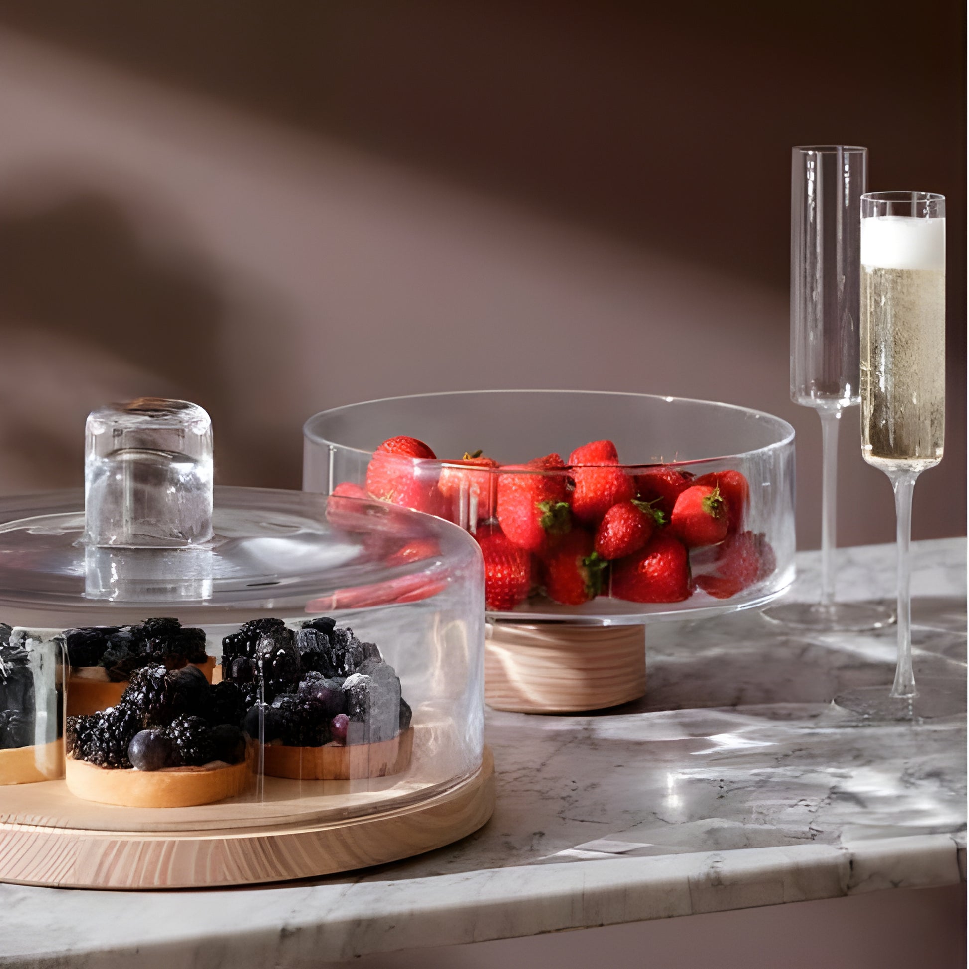 Marble table with Lotta pedestal bowls holding strawberries and berry tarts, beside flutes of champagne.