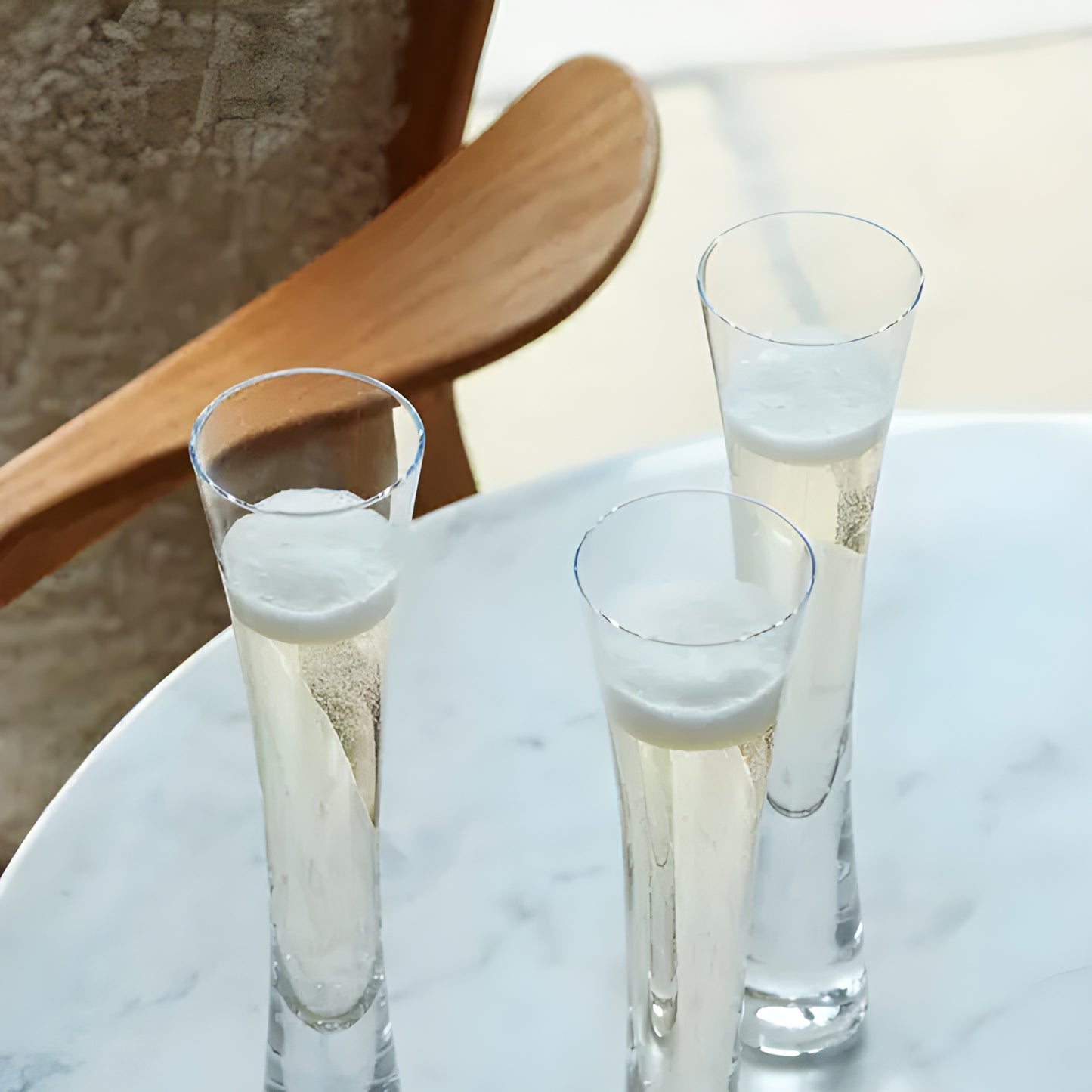 Three tall champagne flutes on a marble table with wooden chair in background.