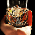 Hand holding a metal-inlaid whisky glass with stag design and ice cubes.
